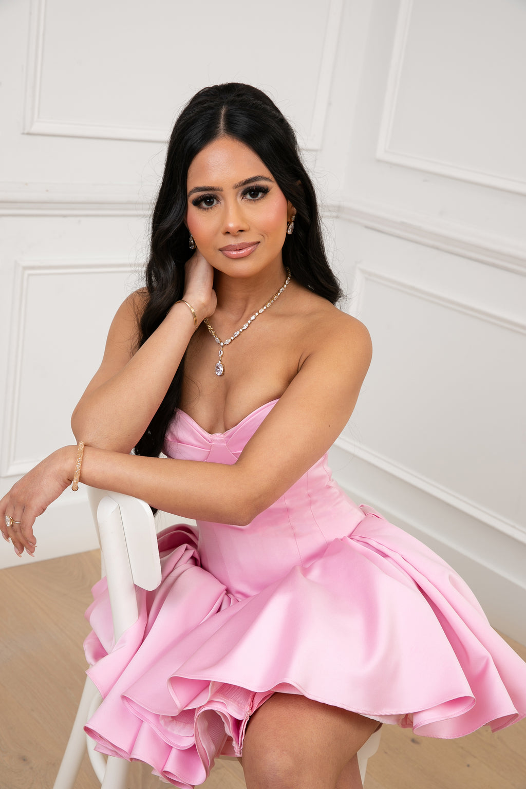 Model wearing The Palais Dress by Brunell by Shar, seated in a pink strapless satin mini dress with sweetheart neckline.
